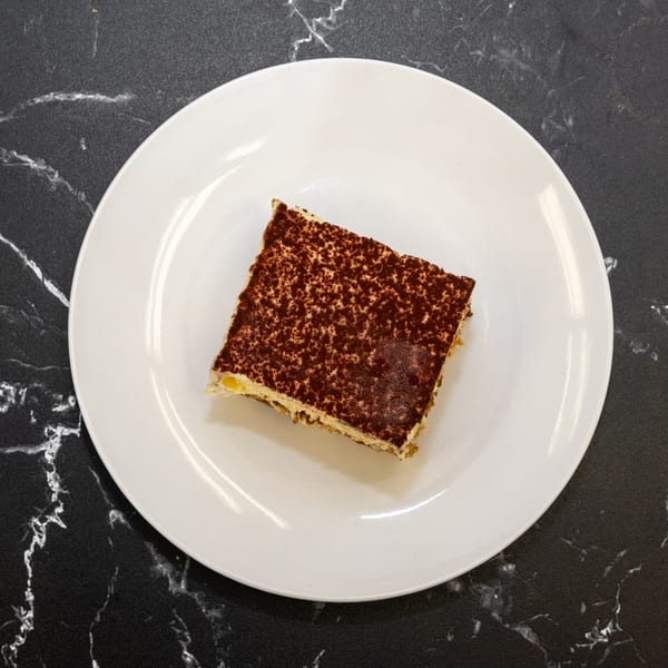 A slice of tiramisu with cocoa powder dusting on a white plate, on a black marble surface.