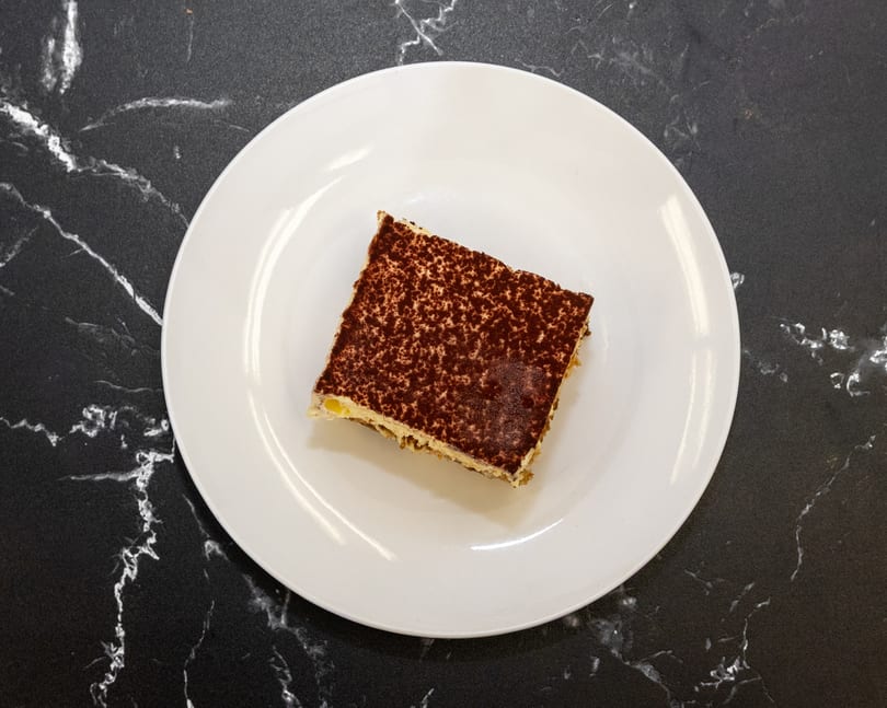 A slice of tiramisu with cocoa powder dusting on a white plate, on a black marble surface.