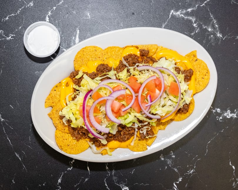 A plate of nachos with ground beef, melted cheese, lettuce, tomatoes, and red onions, with a side of sour cream.