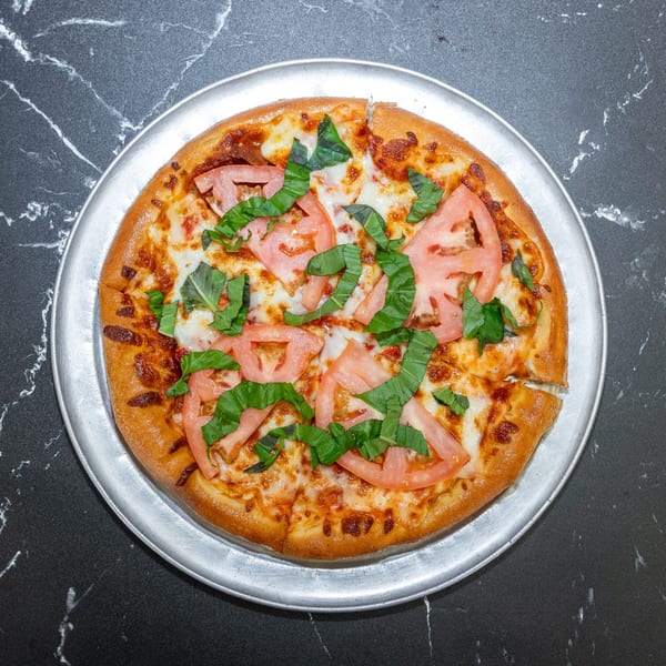 A whole pizza with sliced tomatoes and basil on a metal pan, on a black marble surface.