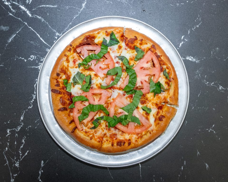 A whole pizza with sliced tomatoes and basil on a metal pan, on a black marble surface.